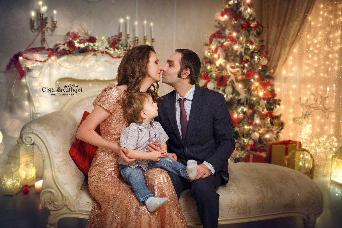 Семейная пара позирует на фоне новогодних огоньков, а их маленький сын сидит у них на коленях.