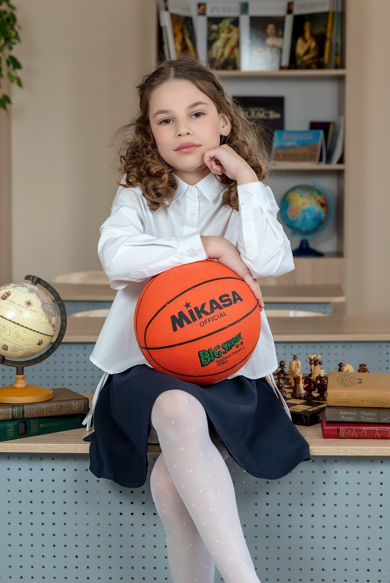 Девочка с ярким баскетбольным мячом сидит на парте позируя профессиональному фотографу.