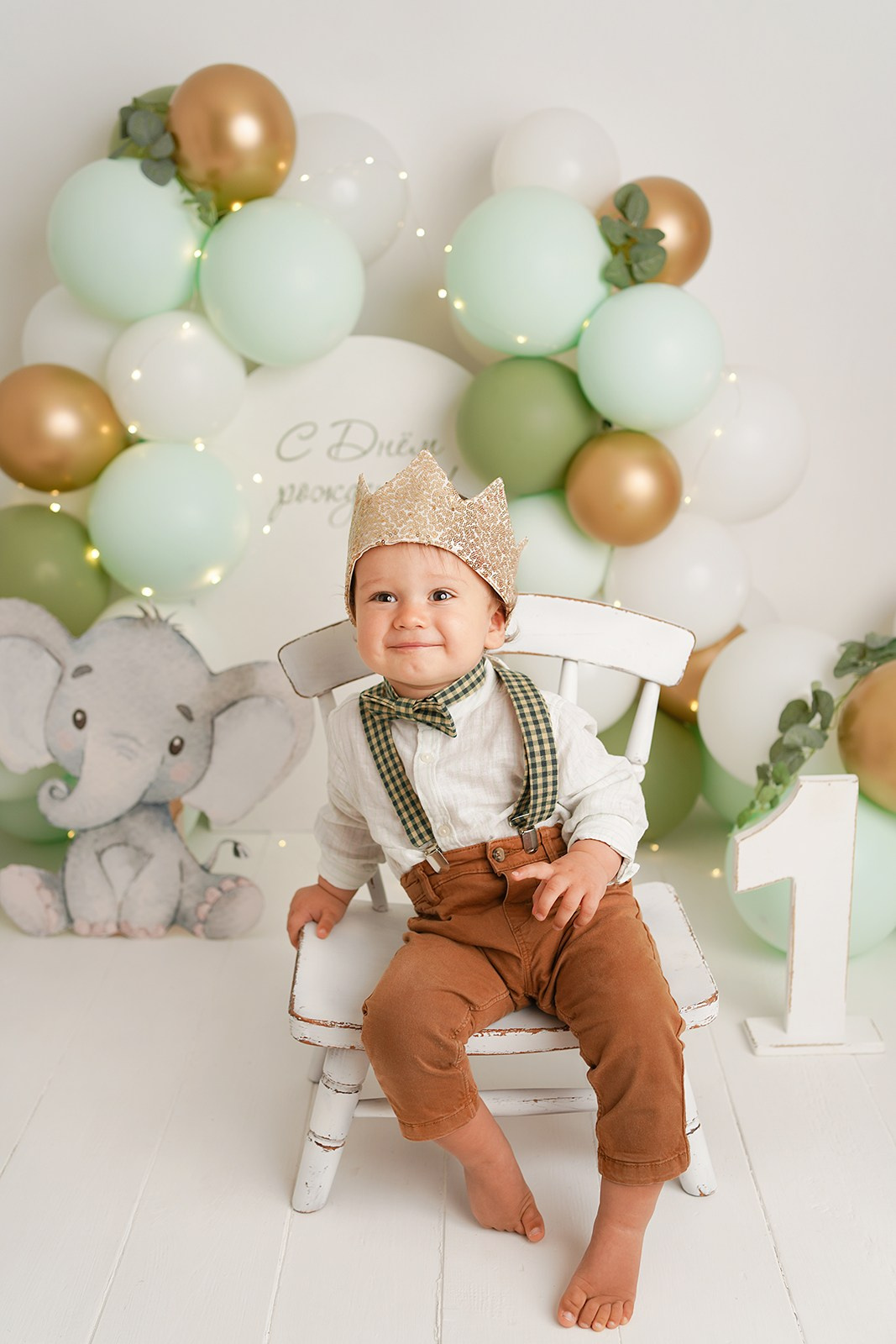Портрет  малыша на день рождения. Он сидит на стульчике в короне в окружении зеленых шариков. огоньков.