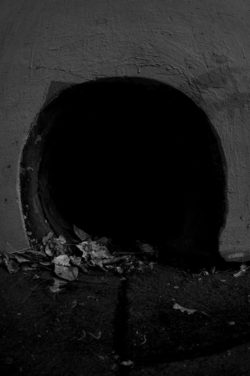 -белая уличная фотография: тёмный круглый проём в грубой стене здания, у основания лежат обломки и камни, резкий контраст света и глубокой тени, минималистичная городская текстура, Вадим Топорёнок, проект Манифест теней, археология душ, городские шрамы, монохромная деталь архитектуры.