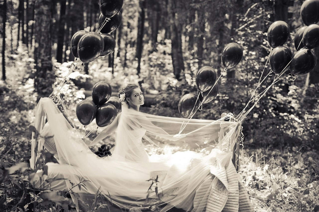 невеста в длинной красивой фате в контровом свете сидит на кровати с привязанными черными шарами  