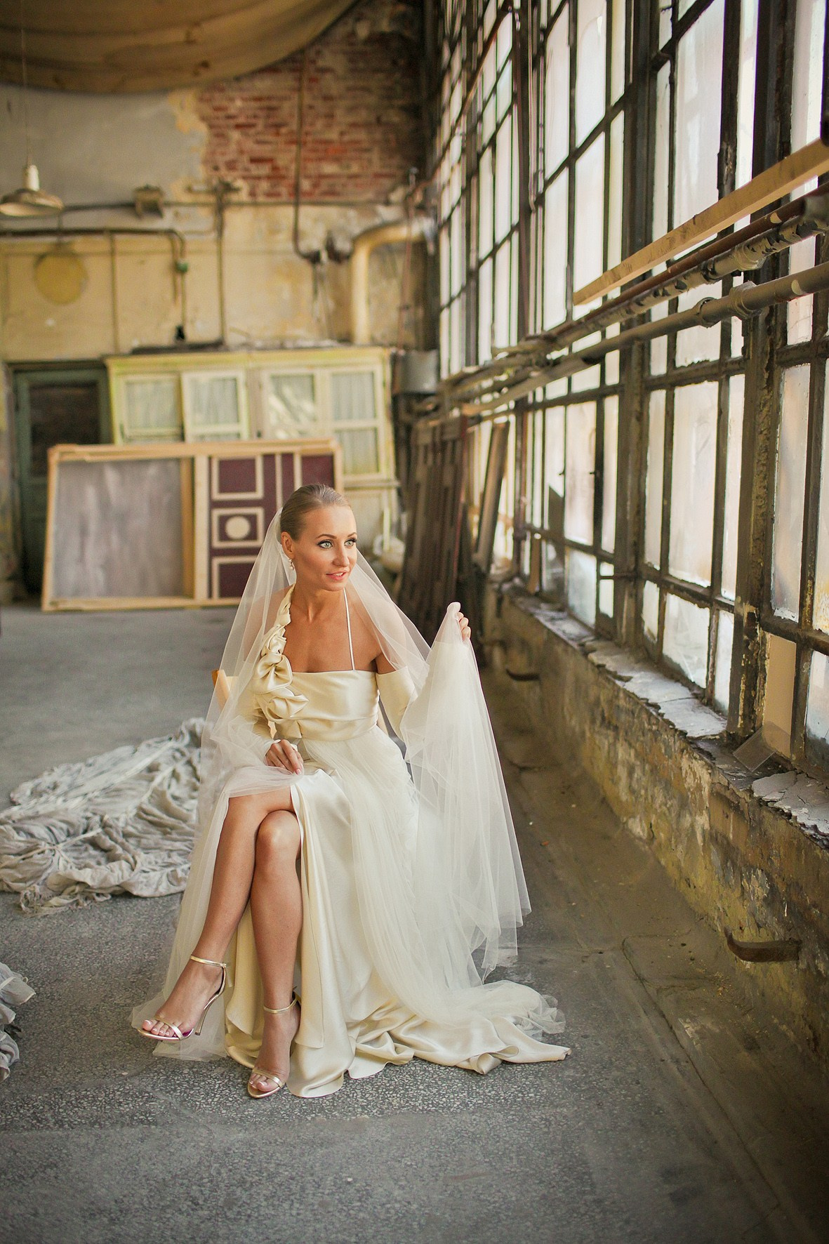 невеста с обнаженными ногами в длинной фате позирует на фоне больших окон в мастерской художника 