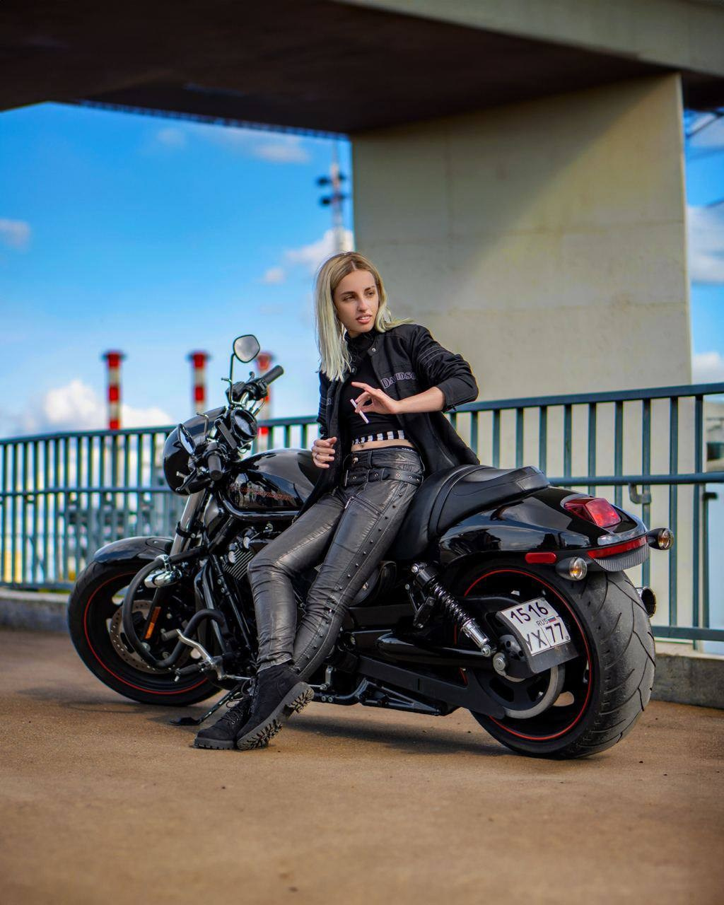 девушка байкер стоит около своего мотоцикла в черном кожаном образе и курит