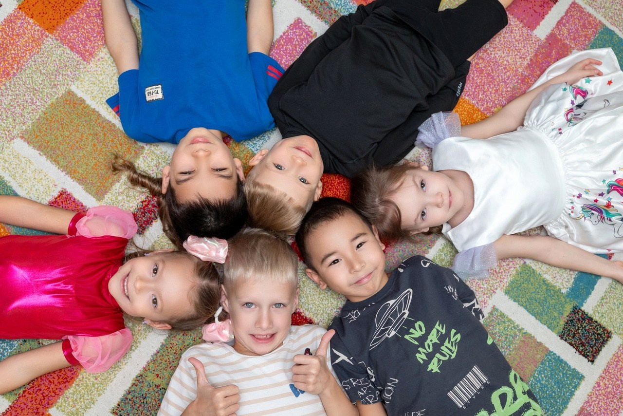 Группа детского сада позирует фотографу лёжа на полу голова к голове.