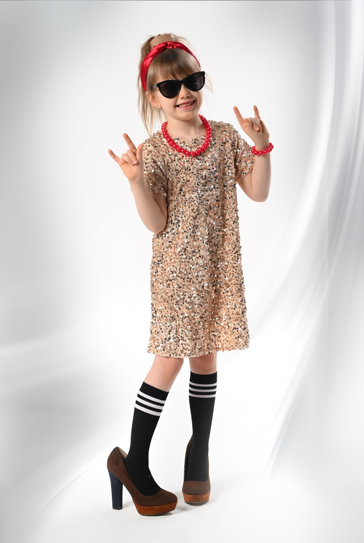 Девочка в платье очках и больших туфлях показывает жест всем мир в фотостудии