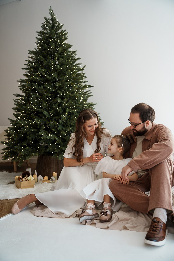 Семейная новогодняя фотосессия в студии Новосибирска: мама, папа и дочка 5 лет смотрят друг на друга с улыбкой на фоне елки. Фотограф Едапина Дарья.