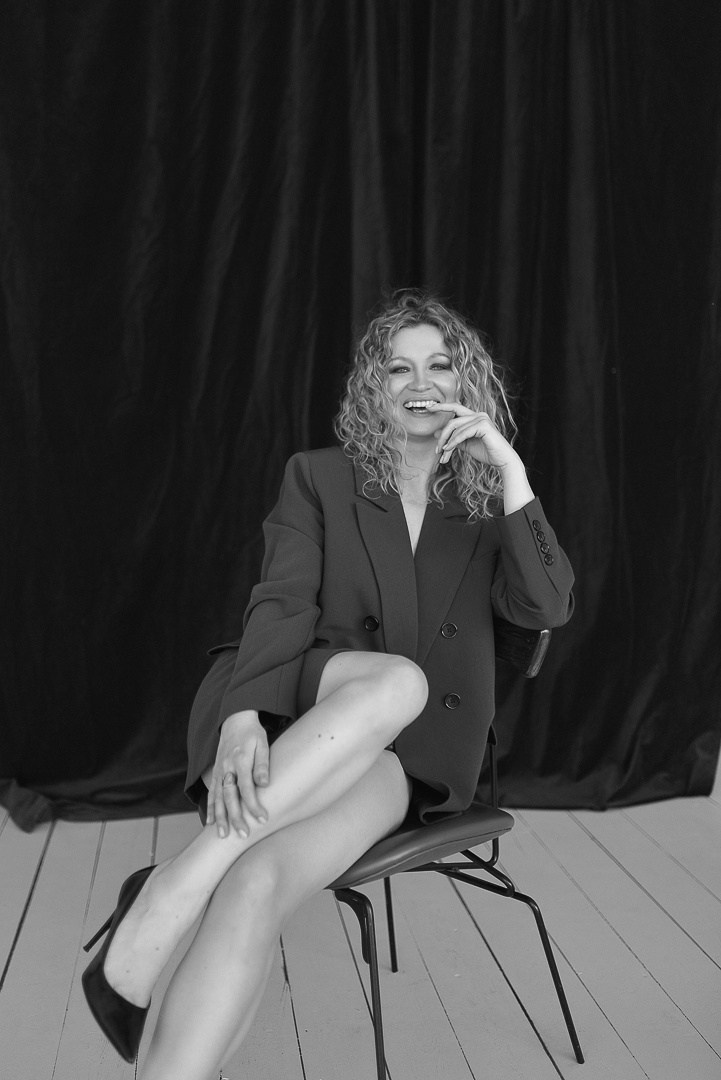 Черно белая фотография девушки в пиджаке и белье с красными туфлями, которая сидит на стуле на черном фоне.