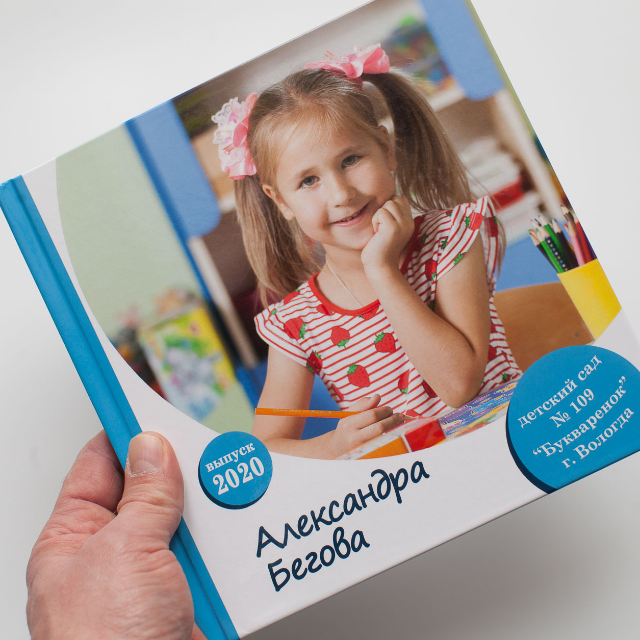 Рука держит квадратную выпускную фотокнигу с девочкой на обложке.