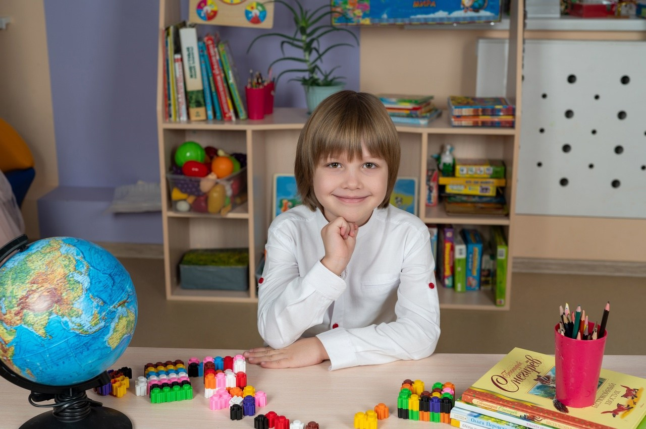 Симпатичный мальчик сидит за столиком рядом с глобусом и фотографируется на выпускной альбом.