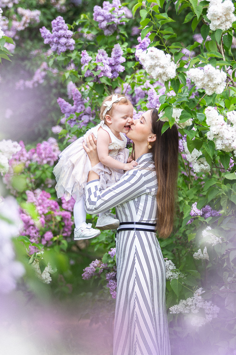 Мама с доченькой малышкой в сиреневом саду улыбаются и целуются.