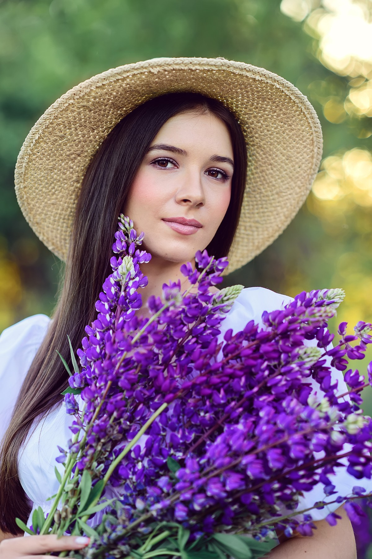 молодая красивая девушка в соломенной шляпке держит в руках букет фиолетовых люпинов