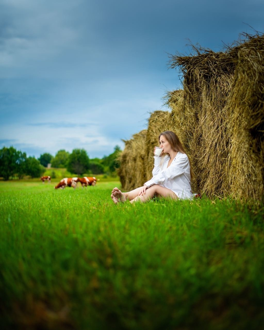 девушка в белой рубашке  в деревне пьет молоко, сидя около стога сена