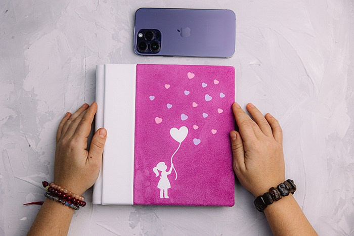 Вид сверху: руки фотографа лежат на фотокниге из розовой замши. Рядом лежит iPhone для понимания масштаба изделия. Лаконичный современный дизайн.