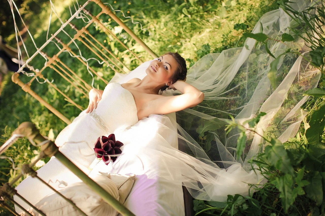 невеста в белом платье с фатой на земле лежит на старинной кровати на фоне зеленой  поляны 