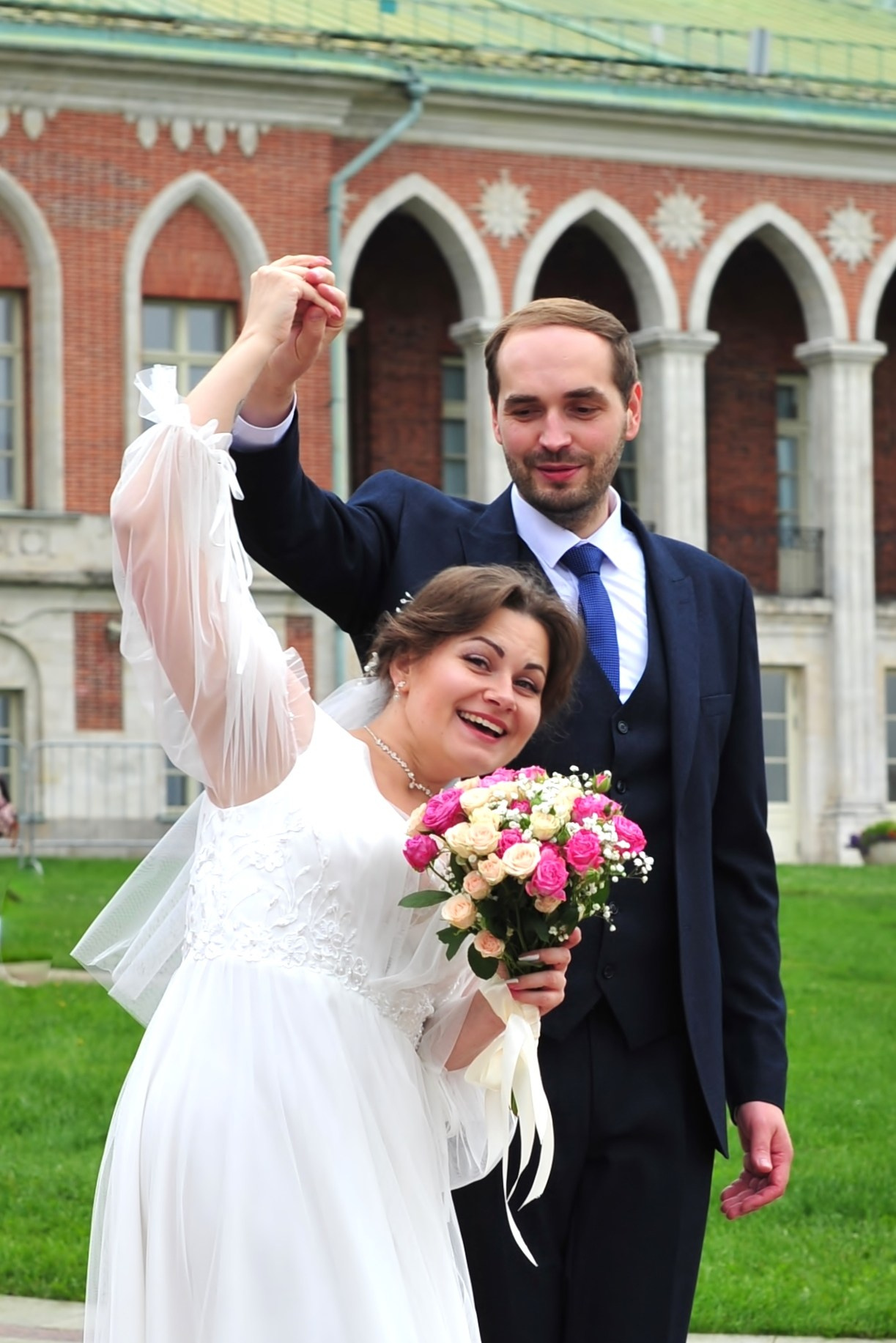 Свадьба в Царицыно пасмурным летним днем, новобрачные держатся за руки на фоне дворца из красного кирпича 