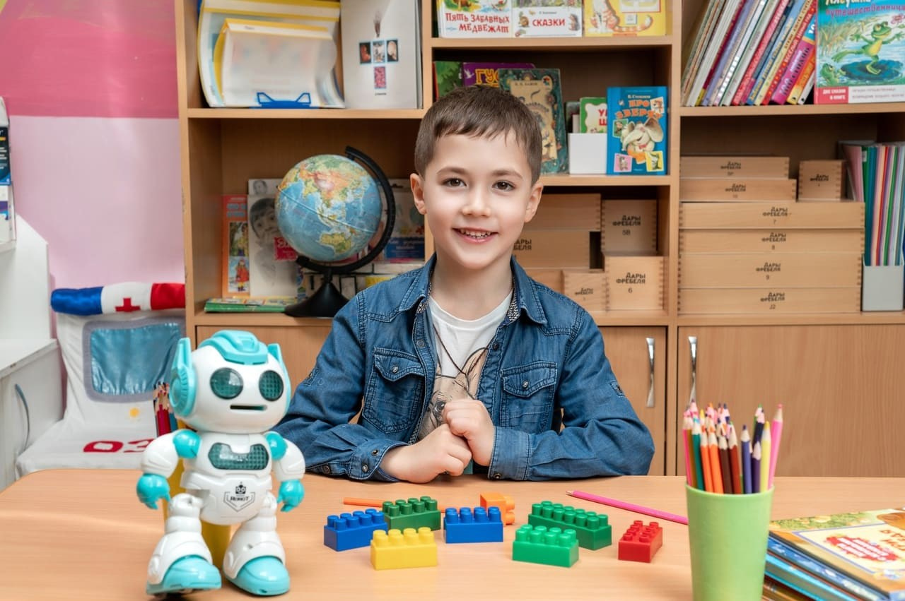 Симпатичный парень выпускной группы детского сада сидит за столиком с роботом.