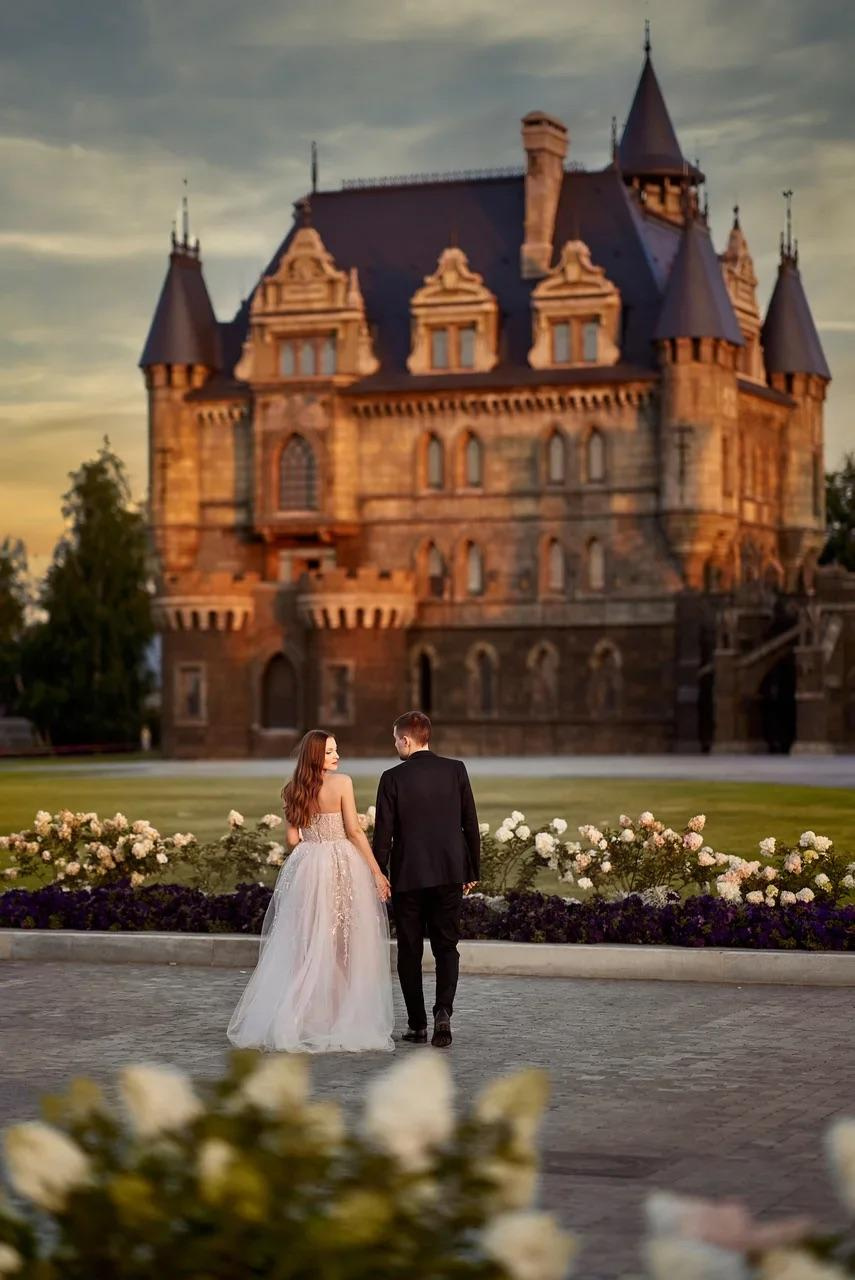 Невеста в белом платье позирует перед главным входом в замок Гарибальди, окружённом цветущими кустами и вечерним светом.