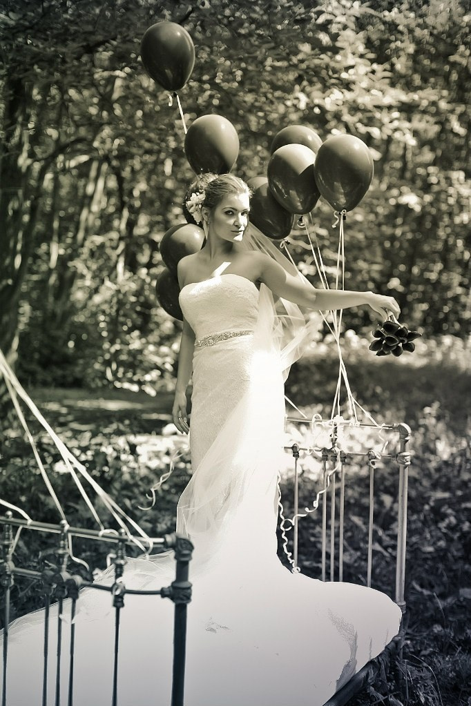 невеста в белоснежном платье посреди леса стоит на старинной кровати украшенной черными воздушными шарами 