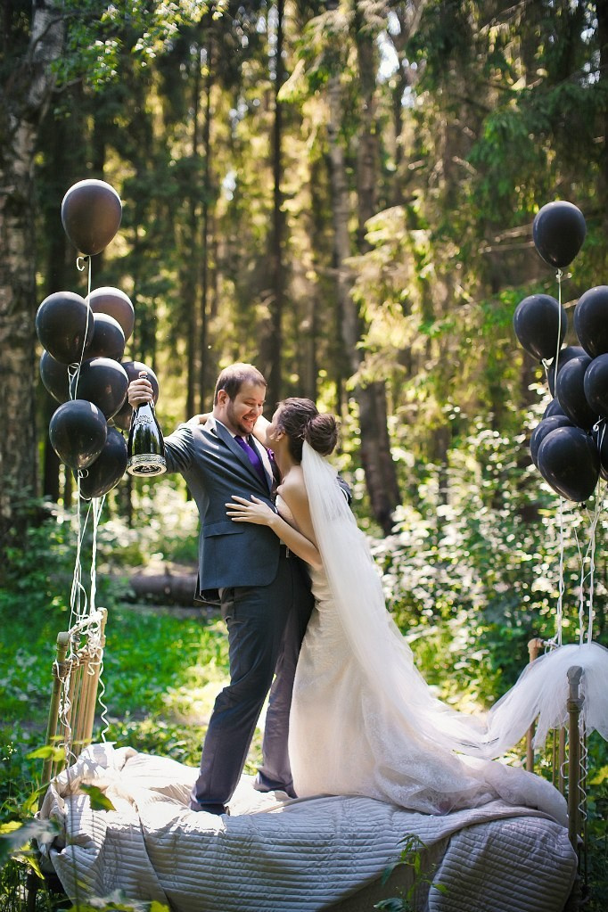 жених с бутылкой шампанского обнимает невесту в белом роскошном платье стоя на кровати в лесу