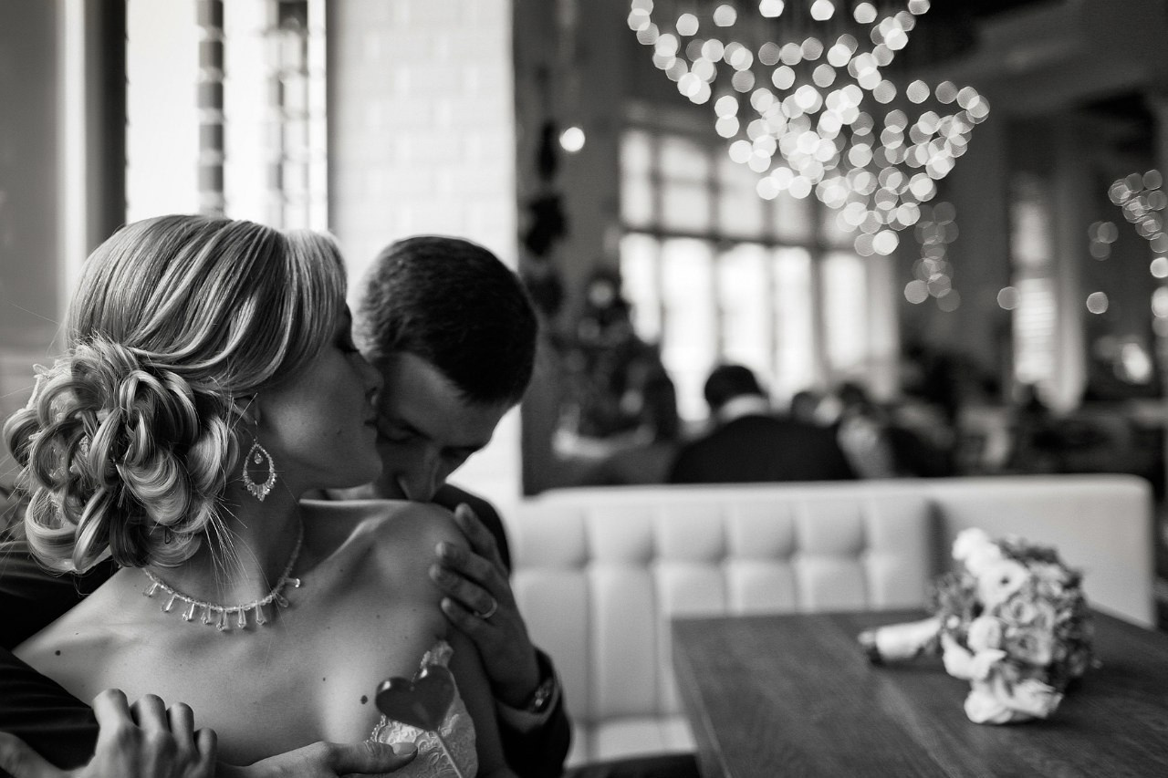 молодожены в роскошном ресторанном зале. жених обнимает и целует невесту в плече 