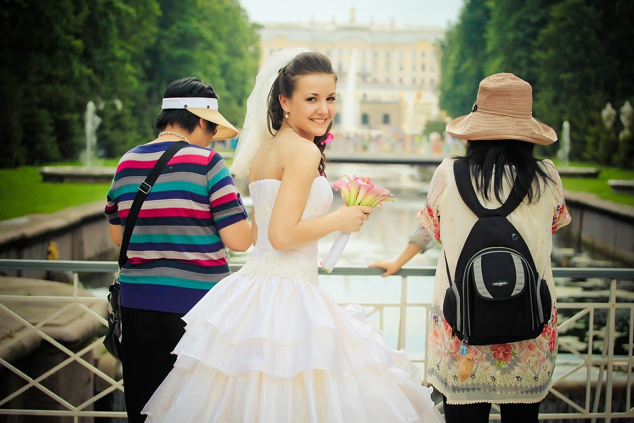невеста в пышном платье позирует на фоне китайских туристов в петергофе