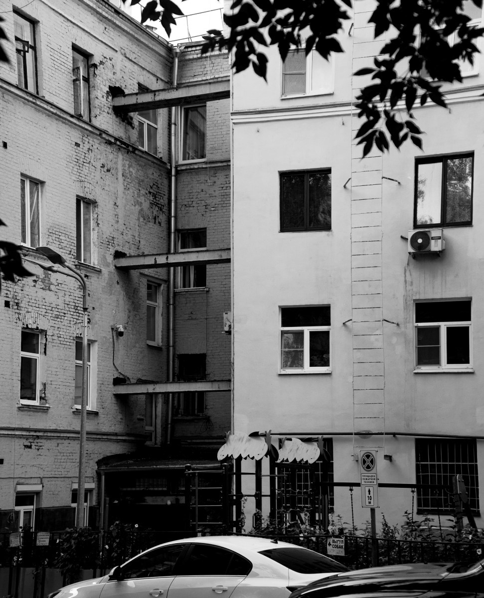 Черно-белая уличная фотография: узкий двор между двумя зданиями, слева обветшалый фасад с обнажённым кирпичом и наружной металлической лестницей, справа светлая стена с контуром замурованной двери и кондиционером, на переднем плане крыша автомобиля, ветви дерева сверху, городская архитектура, Вадим Топорёнок, Манифест теней, геометрия города, текстуры времени, урбанистический пейзаж.
