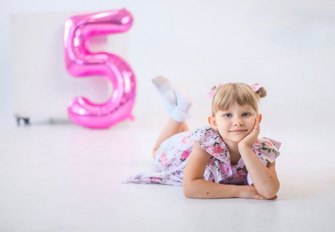Маленькая девочка в цветочном платье лежит на полу, подперев щёку рукой. У неё аккуратные пучки с розовыми бантиками. На фоне — большой розовый шар в виде цифры «5».