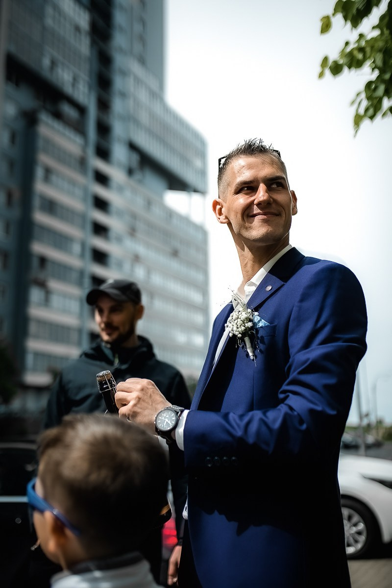 Жених открывает шампанское на фоне ЗАГСа на Васильевском острове в Санкт-Петербурге. Вертикальный поясной кадр.