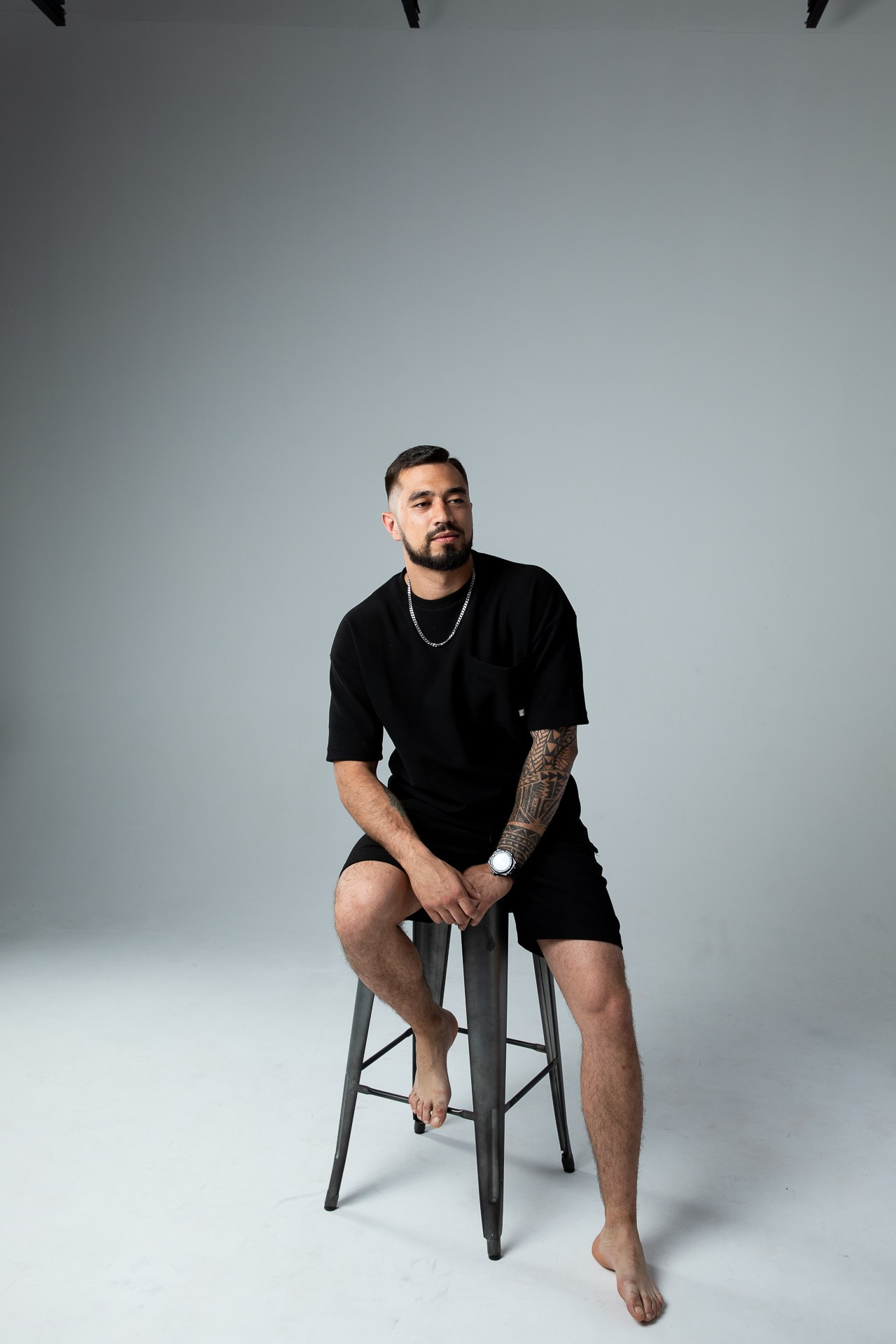 Мужчина брюнет в черной футболке и шортах сидит на высоком барном стуле