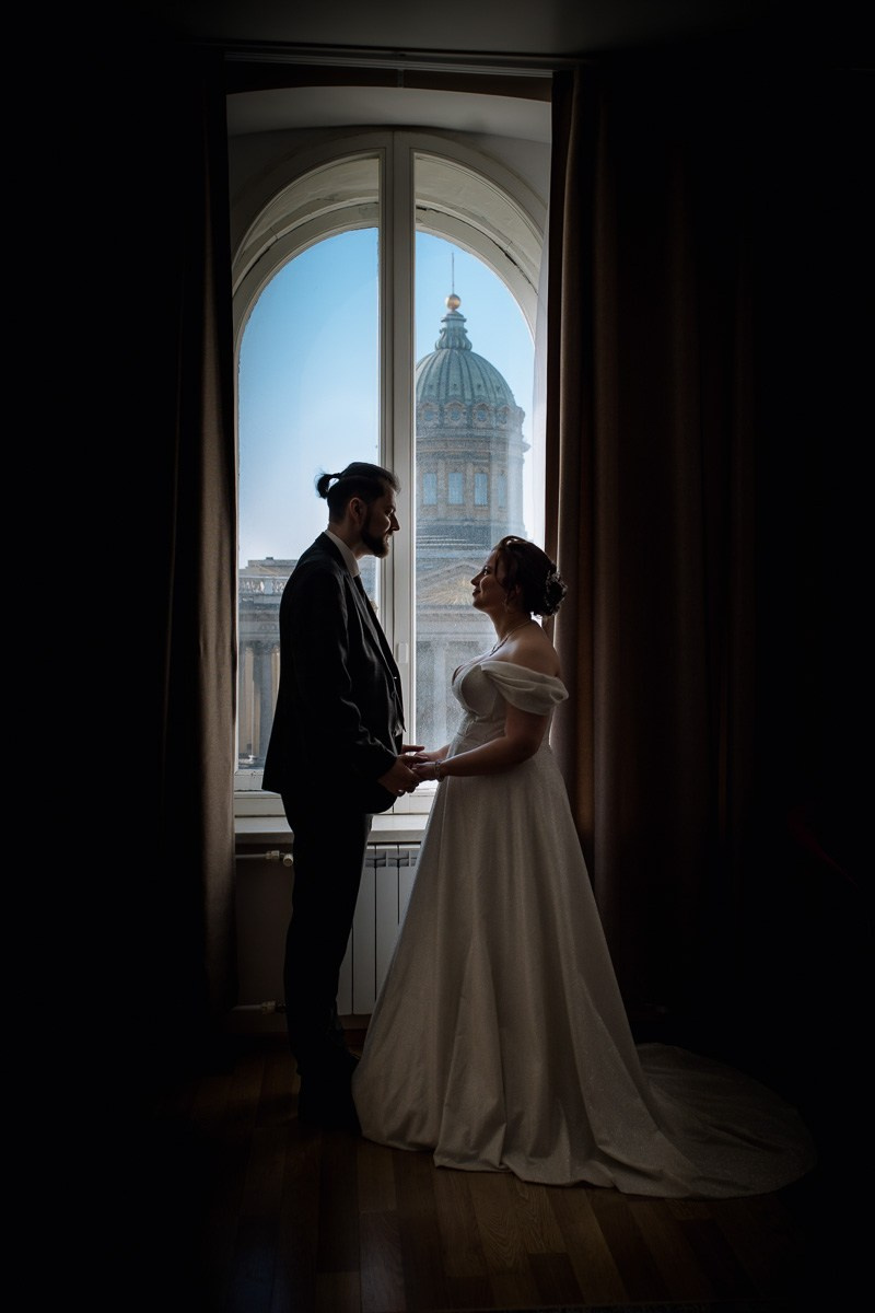 Жених и невеста в отеле смотрят друг на друга и держатся за руки , а за окном Казанский собор Петербурга