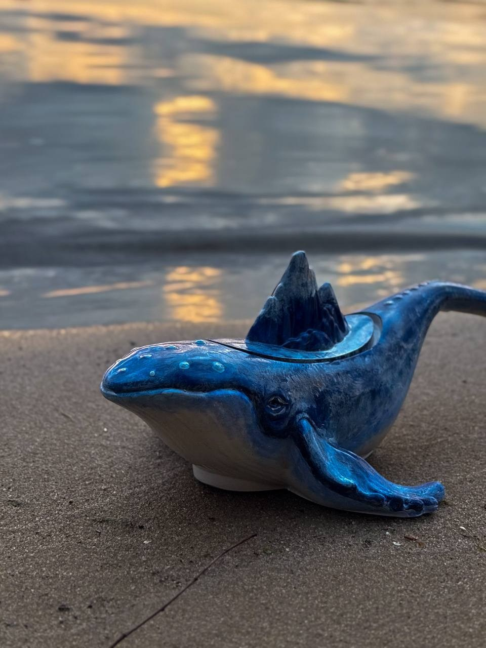 Фото синей свечи-дельфина на песке у воды — предметная съёмка на закате.