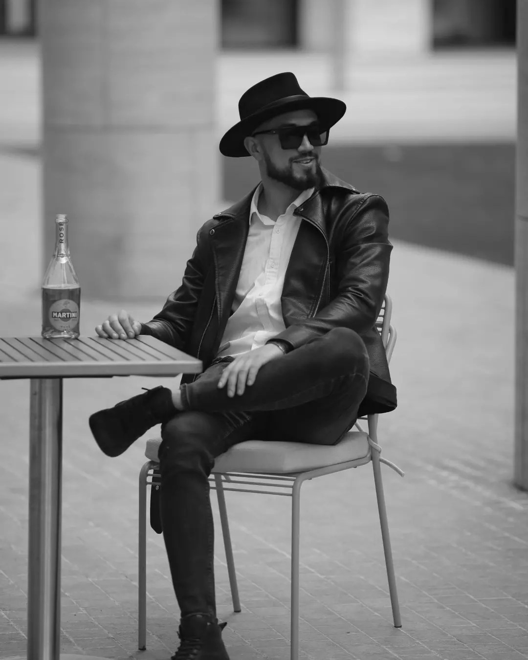 Мужчина с бородой в кожаной куртке, шляпе и солнечных очках сидит за столом с бутылкой вина