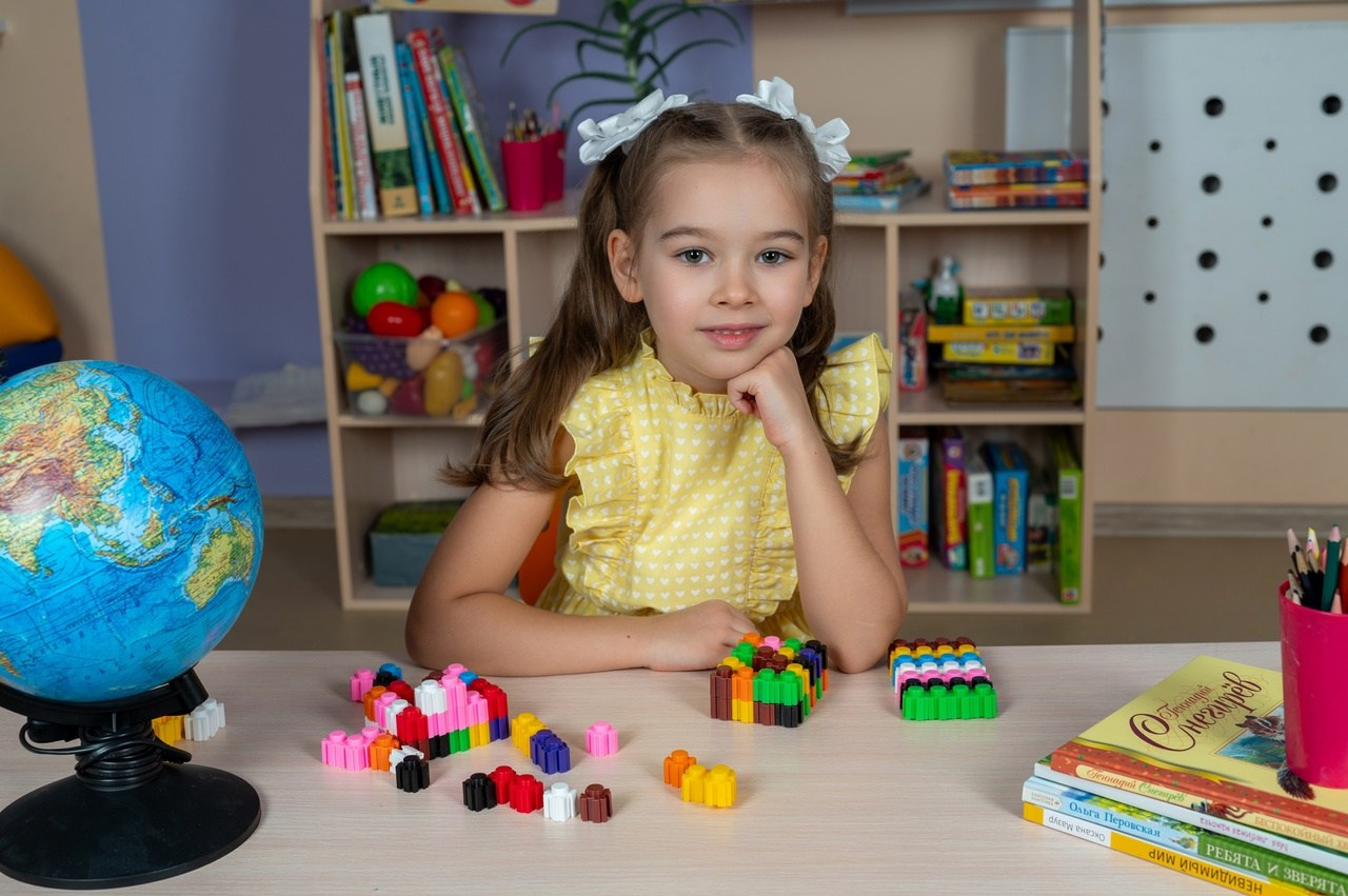 Девочка в желтом платье позирует фотографу в группе детского сада.