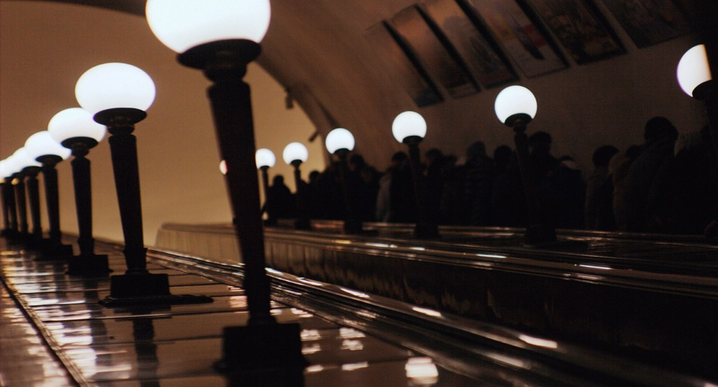 Эскалатор в метро с рядами круглых ламп, силуэты людей наверху, теплый свет, перспектива подземного перехода