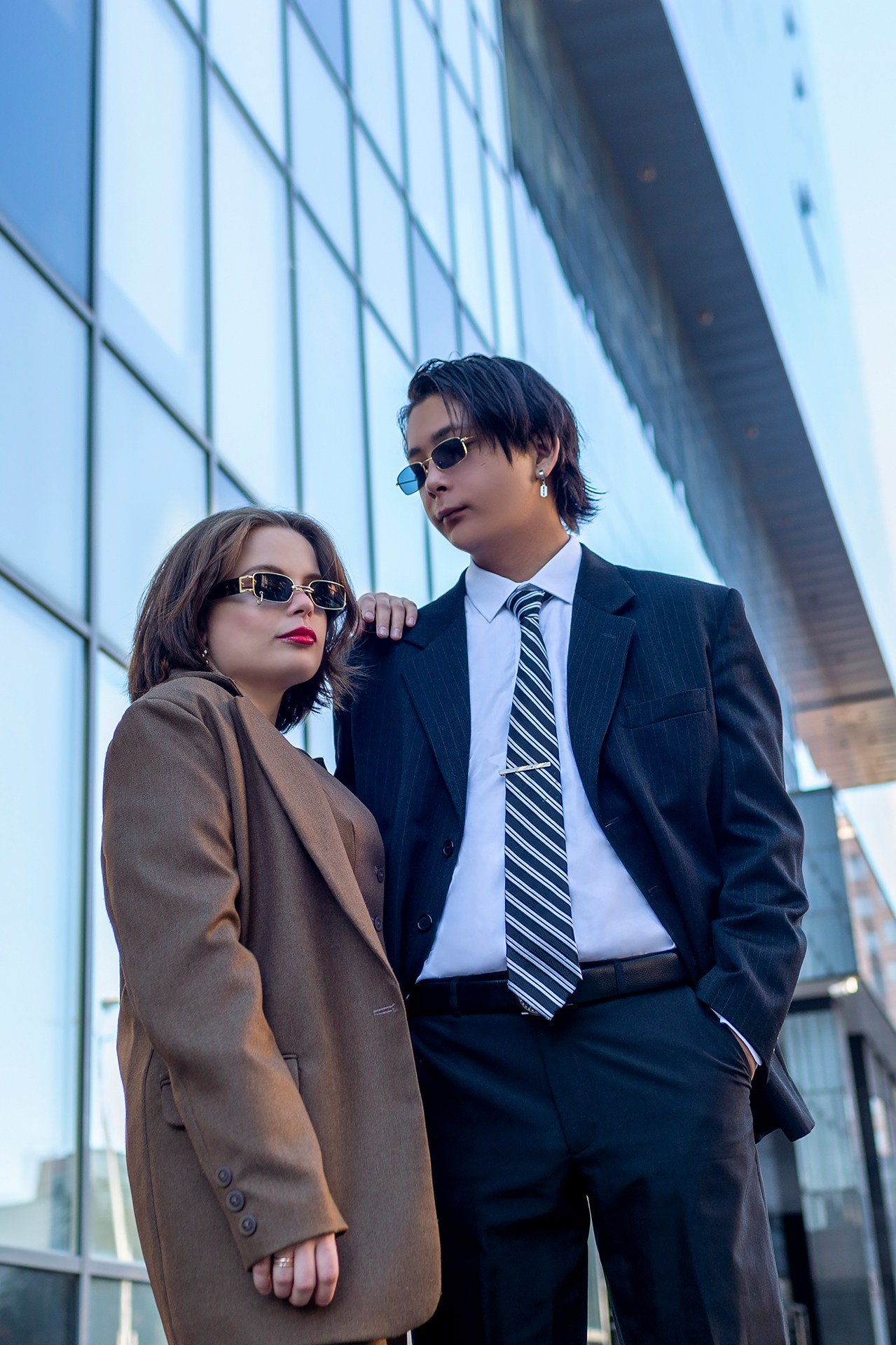 Пара мужчина и женщина в деловых костюмах и темных очках стоят на фоне стеклянного небоскреба.