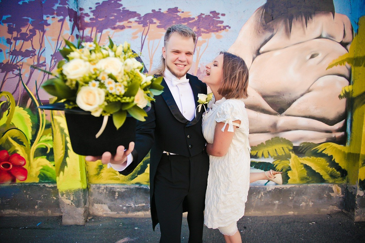 жених в черном фраке на фоне граффити держит в руках шляпу и обнимает смеющуюся невесту