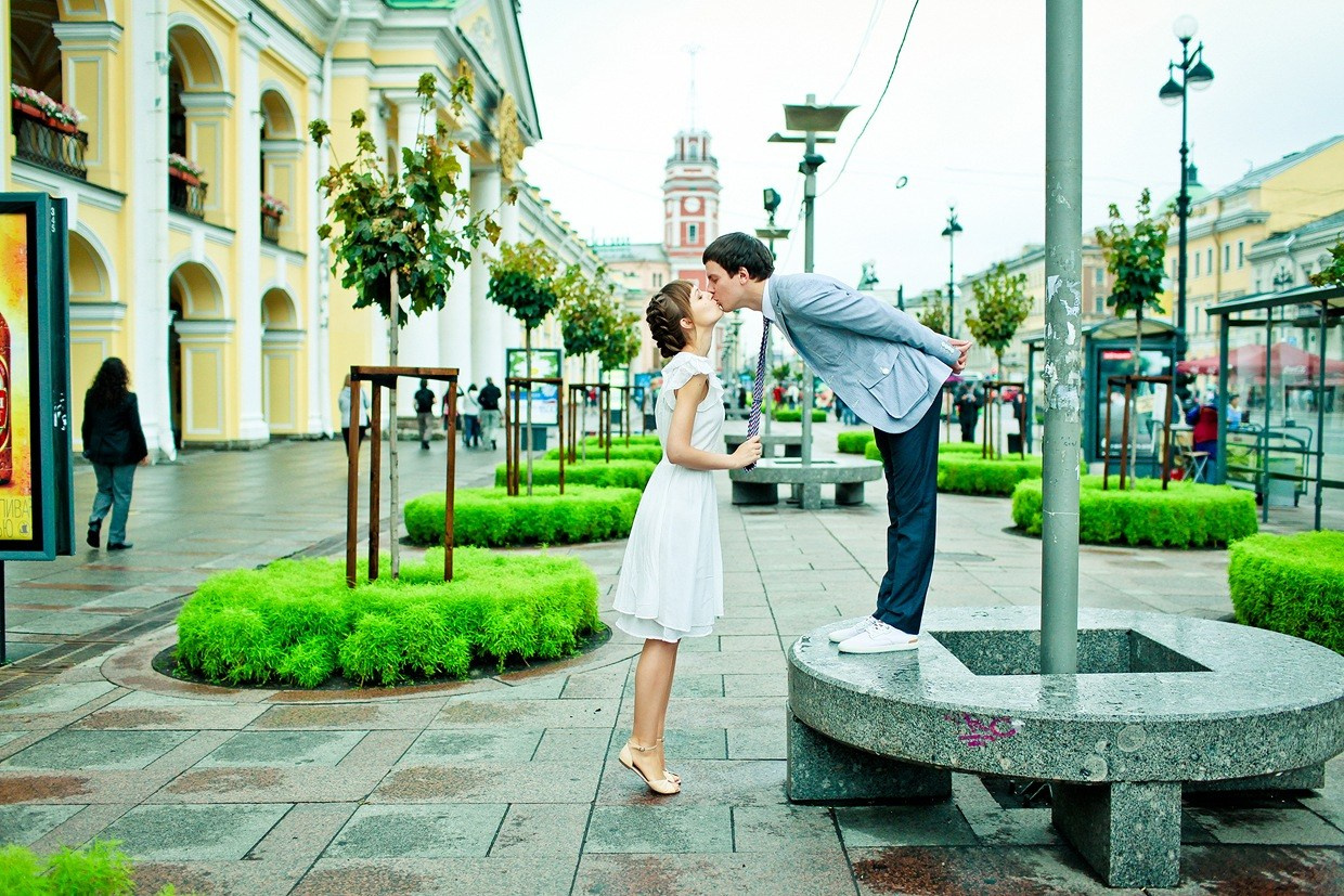 жених в сером пиджаке стоя на каменной скамейке у гостинного двора целует невесту стоящую снизу