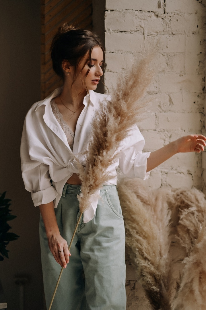 Высокий романтичный  пучок для фотосессии в студиях спб где девушка в белой рубашке стоит у окна с сухими цветами.