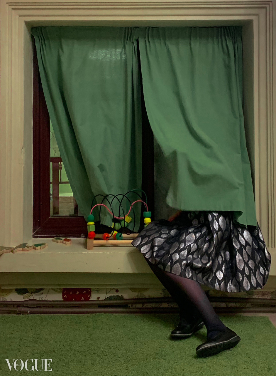 Девочка в праздничном платье спряталась за шторой в окне сл значком Vogue