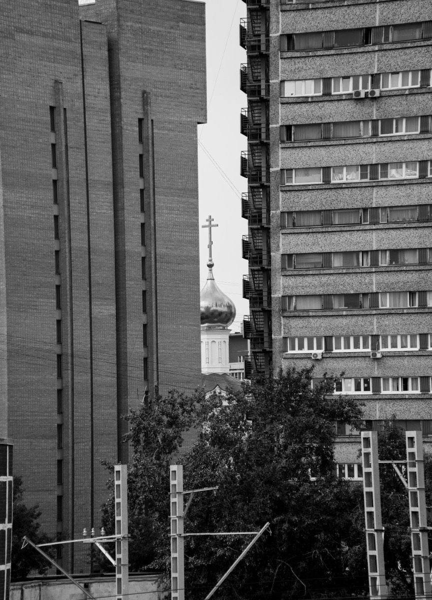 Черно-белая городская фотография: вид между двумя высотными зданиями, в узком вертикальном просвете виден купол православного храма с крестом, на переднем плане кроны деревьев и опоры линий электропередач, контраст урбанистической застройки и сакрального символа, уличная фотография Москвы, Вадим Топорёнок, Манифест теней, археология душ, геометрия города, скрытая память.