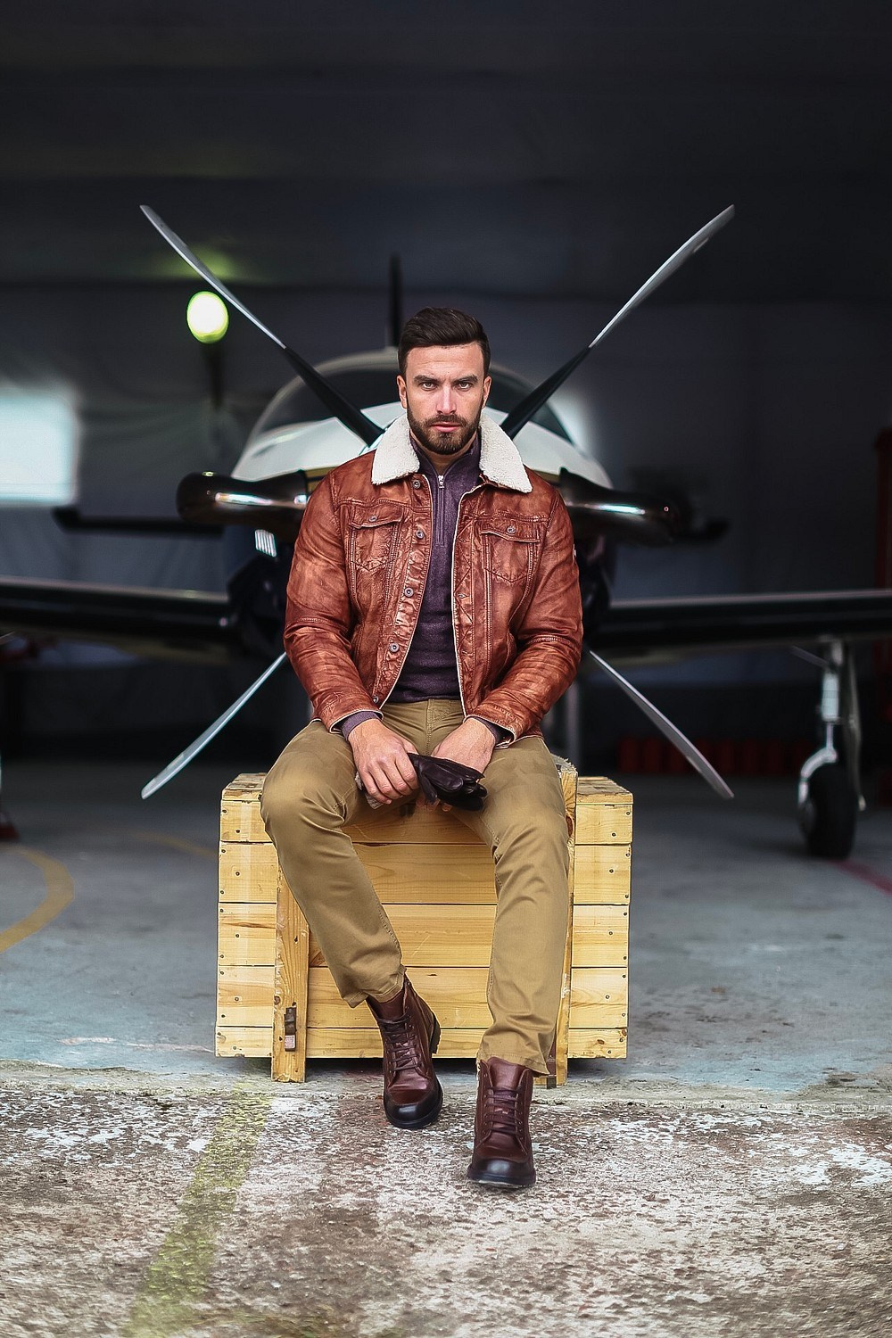 Мужчина с бородой в коричневой кожаной куртке сидит на деревянном ящике на фоне самолета