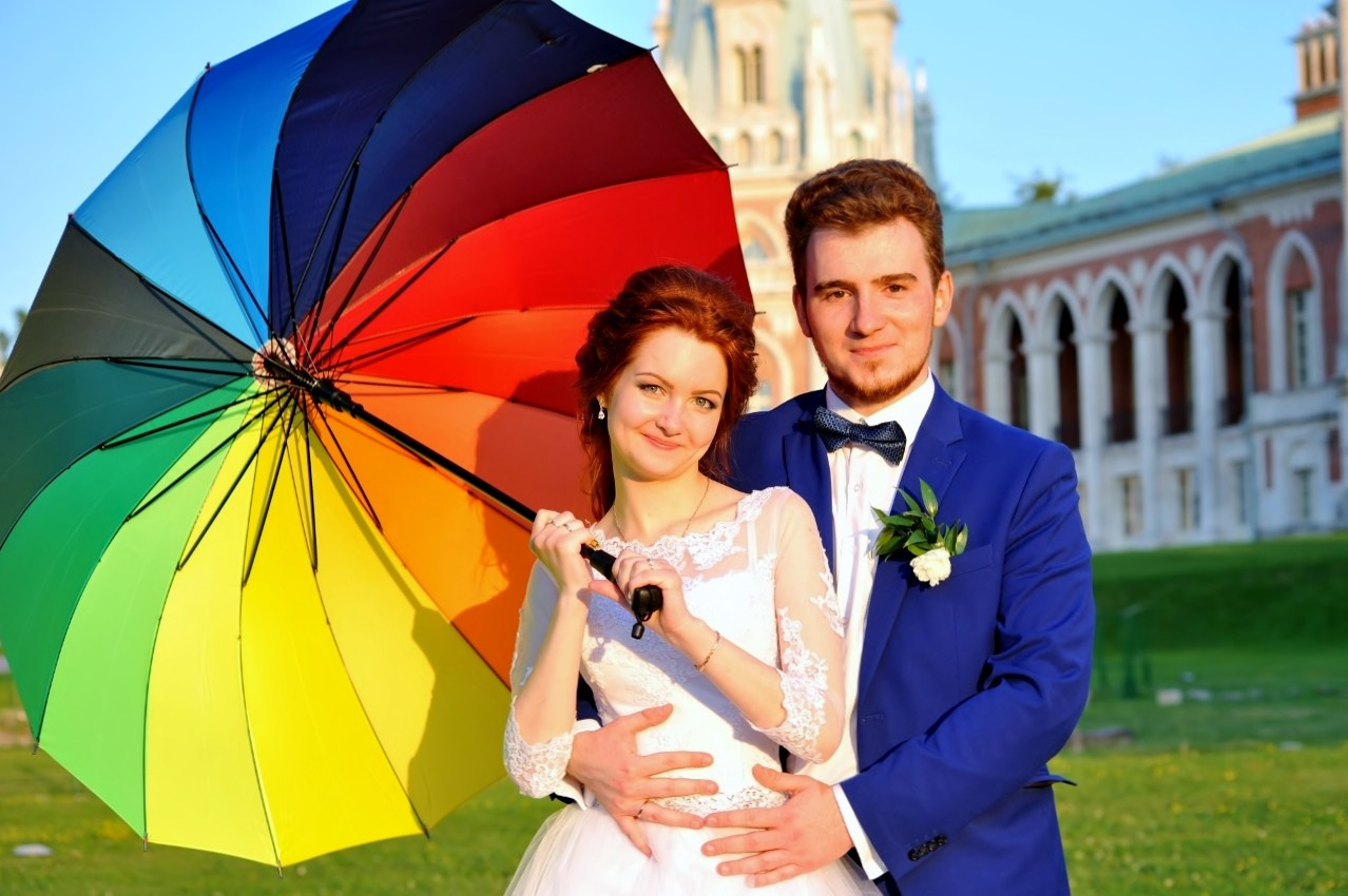Свадебная фотосъёмка в Царицыно летом на закате, молодожены стоят на фоне красно-белого дворца, невеста держит в руках яркий разноцветный зонт от солнца