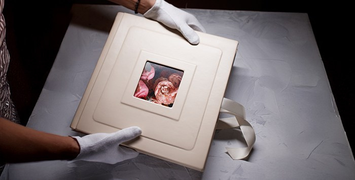 Процесс презентации премиальной фотокниги: руки мастера в белых хлопковых перчатках бережно держат альбом в обложке из бежевой кожи с квадратным окошком и шелковой лентой.