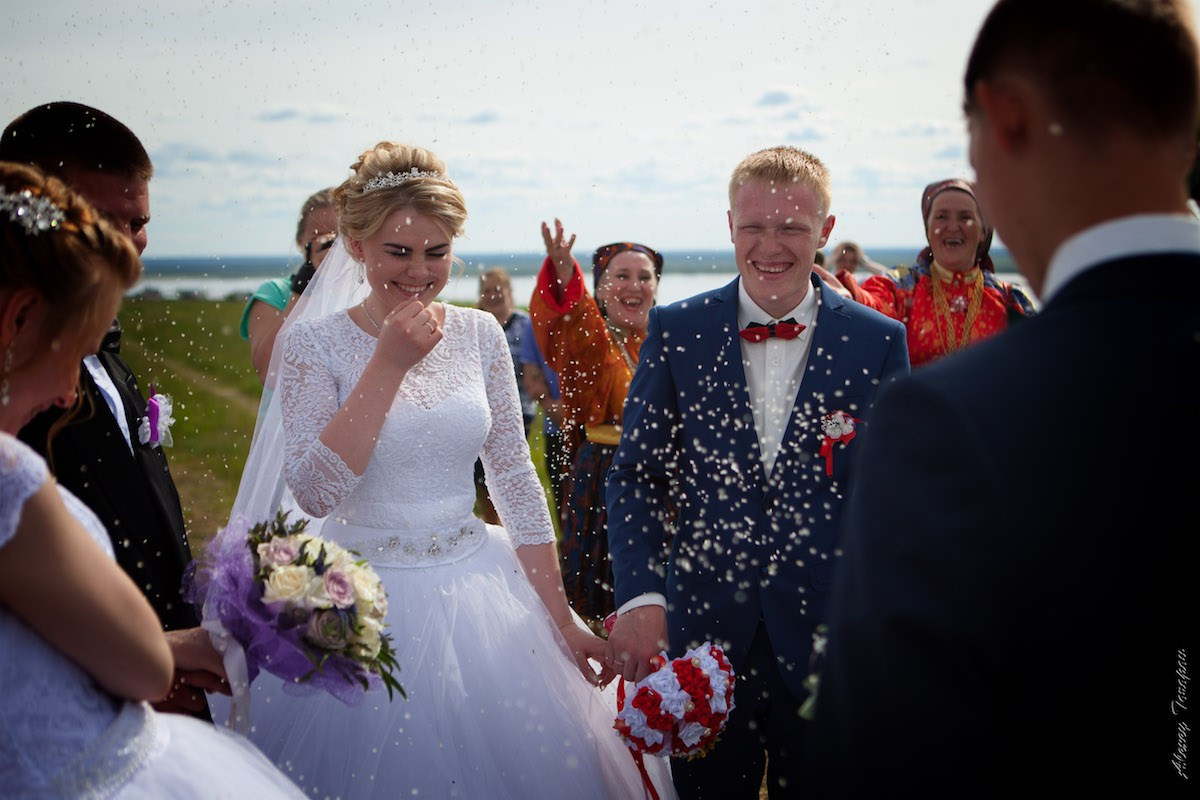 Свадебная пара на улице: невеста в белом платье с букетом и жених в синем костюме улыбаются, пока гости вокруг осыпают их рисом на фоне неба и воды.