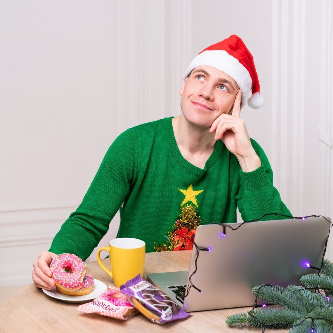 Парень в зеленом свитере и новогодней шапке сидит у ноутбука с чашкой и пончиком