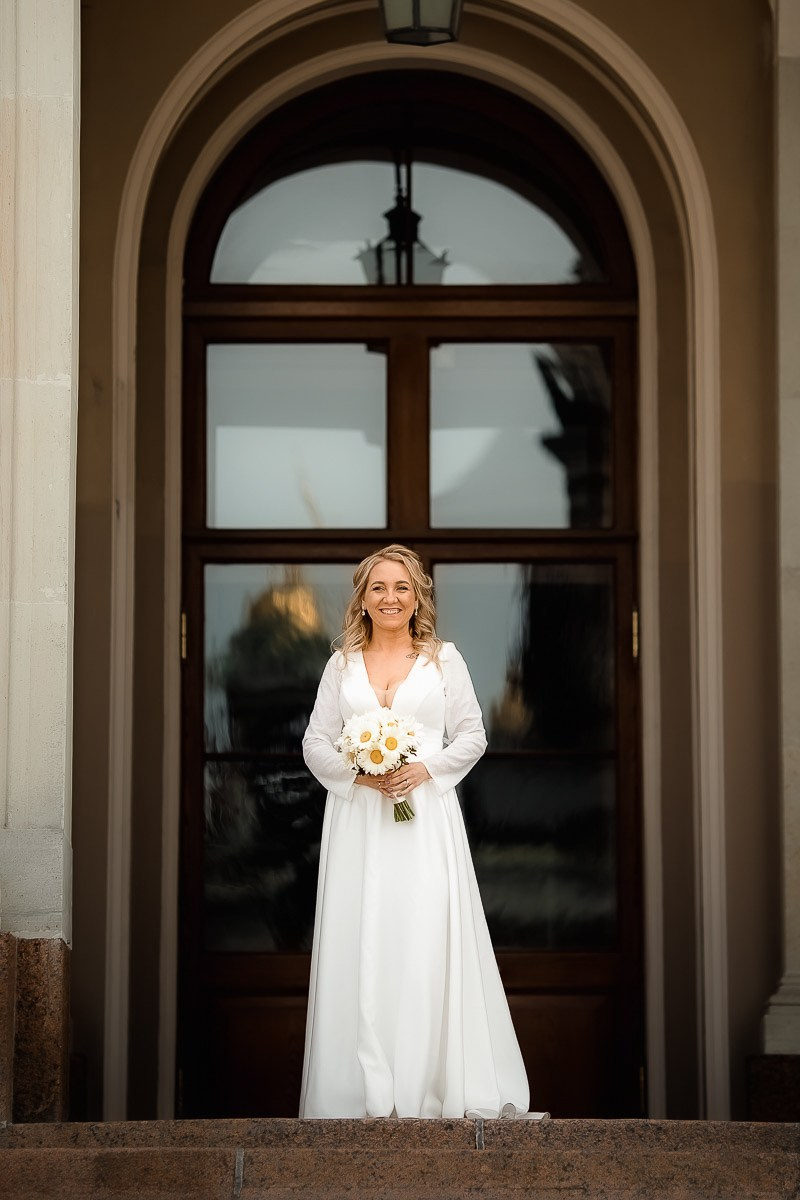 невеста смотрит в кадр, стоя в арке на крыльце заксобрания в Санкт-Петербурге. Вертикальный ростовой кадр.