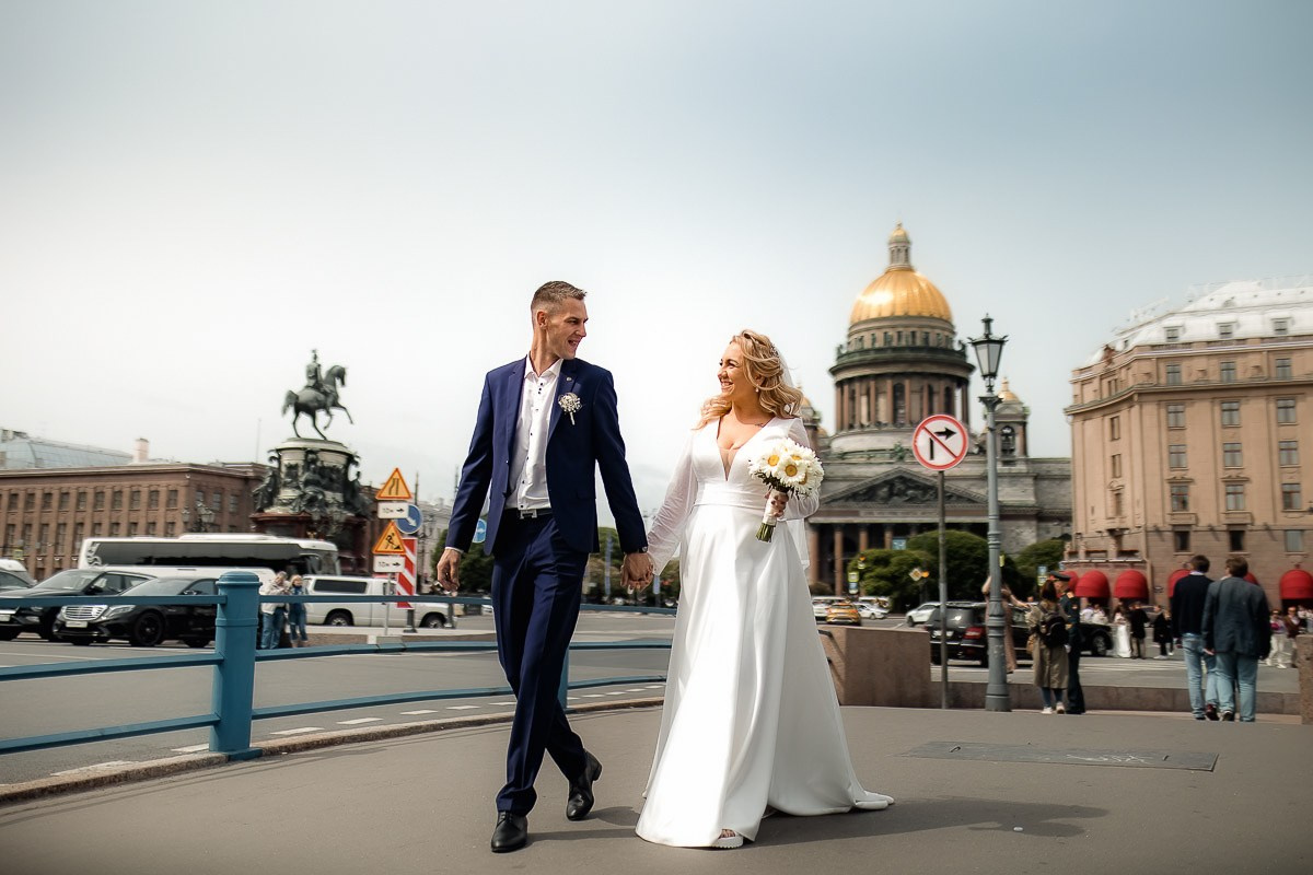 Жених и невеста идут днём по улице за руку, смотрят друг на друга на фоне Исаакиевского собора в Санкт-Петербурге