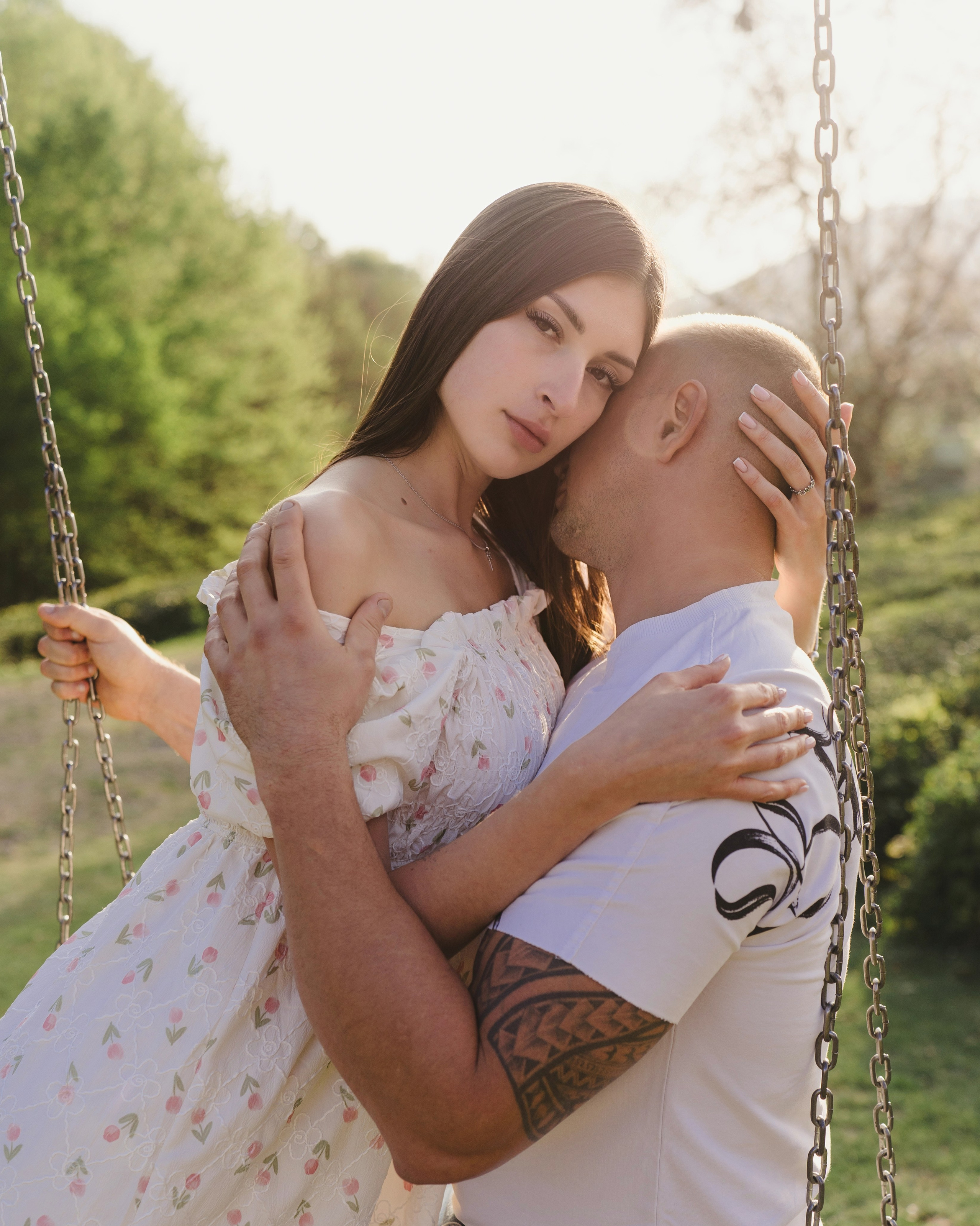 На фотографии изображена пара, обнимающаяся, сидя на качелях. Женщина одета в цветочное платье, а мужчина с лысой головой, в белой футболке с черным рукавом, на котором изображены узоры, напоминающие племенные или маори татуировки, на левой руке. Они находятся в окружении природы, что намекает на парк или сад, и видно, что погода ясная и солнечная. На изображении отсутствует текст.