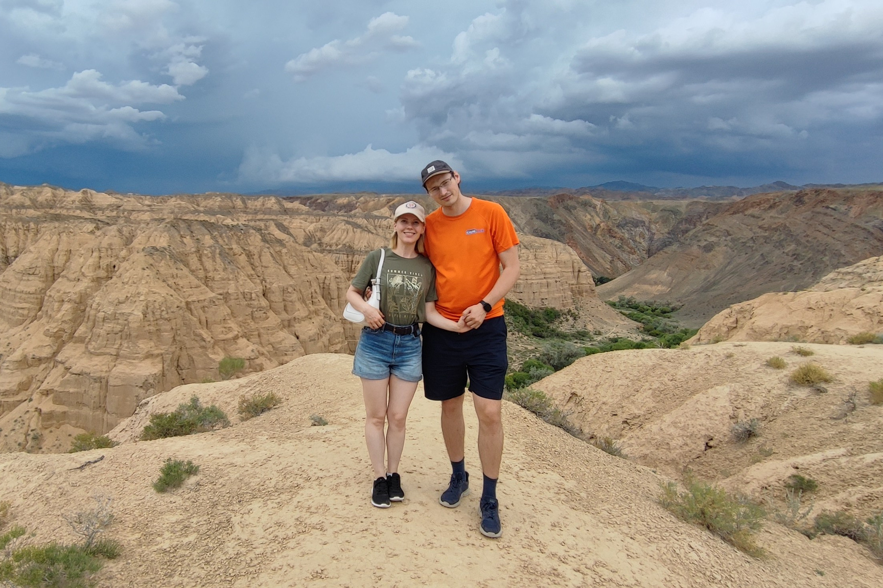 Мужчина и женщина стоят вместе на вершине скалы с видом на пустынный Лунный каньон. Женщина в зелёной футболке, джинсовых шортах и белой кепке, мужчина — в оранжевой футболке, чёрных шортах и кепке. На заднем плане видны скалистые холмы, тёмно-зелёная растительность в долине и драматичное небо с грозовыми облаками, создающее контраст с сухой, песчаной землёй.