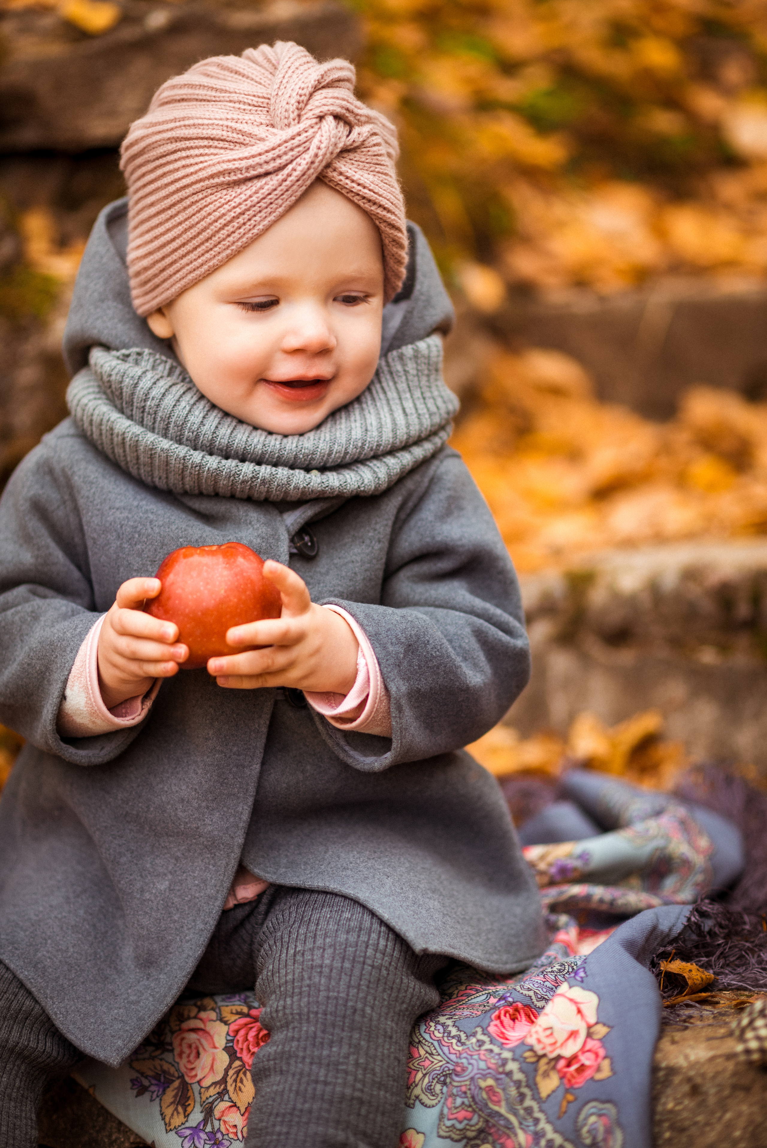 девочка в сером пальто сидит в парке на фоне желтых листьев с красным яблоком в руках